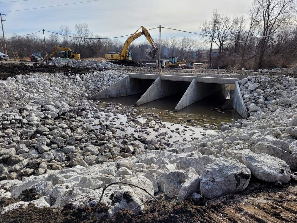 A newly constructed triple-cell concrete box culvert with flared wingwalls and a rock apron, water flowing through it.