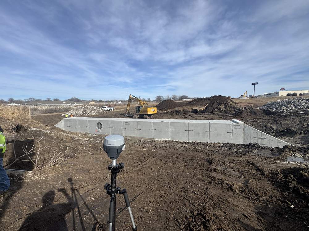 A GNSS survey receiver on a tripod in the foreground of an active job, with a precast concrete box culvert being set behind it and an excavator working beyond that.