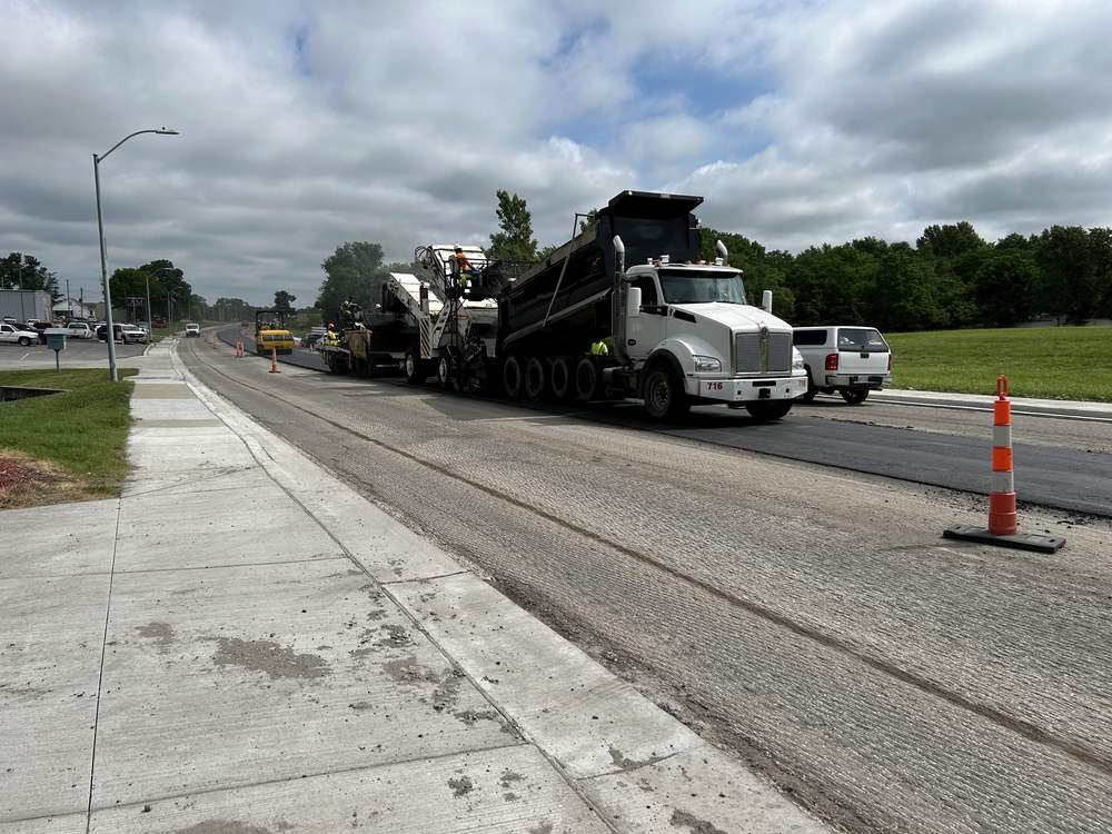 An asphalt paving train laying a mat beside a new concrete curb and sidewalk on an arterial street.