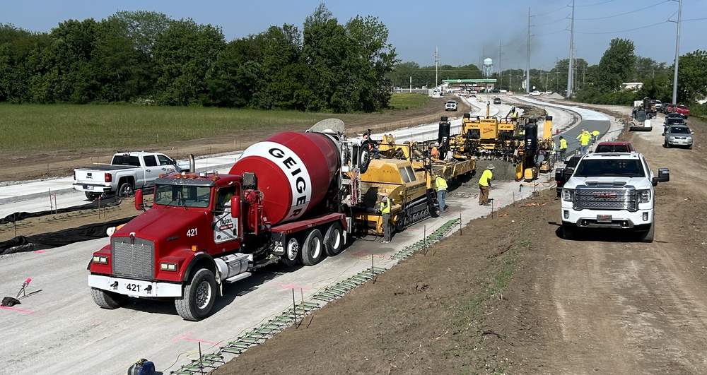 A slipform paver placing concrete pavement on a new public roadway, with a ready-mix truck feeding a belt placer and a crew working alongside.