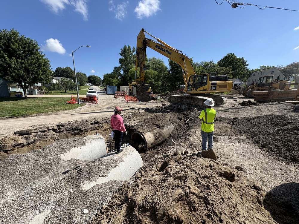 Storm sewer under construction on a city street, the old pipe out, new structures staged along the trench and one lane kept open to traffic.